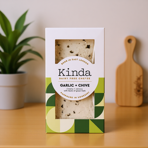 Kinda Garlic + Chive dairy-free cheese packaging on a wooden surface with a plant and cutting board in the background.